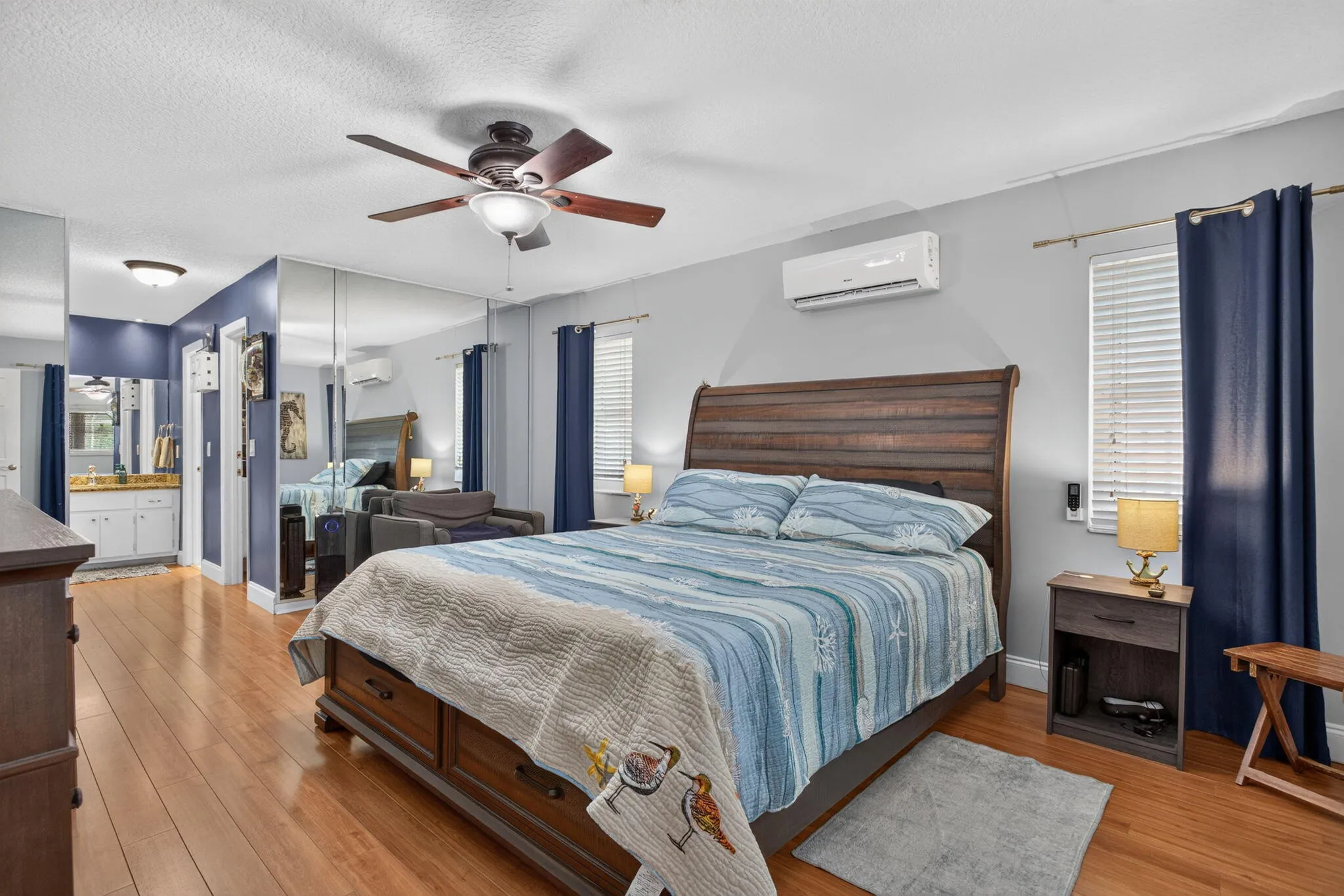 Wood-finish flooring, ceiling fan with integrated lighting, wall-mounted air conditioning unit, mirrored wall, and built-in vanity with white cabinetry and a rectangular sink