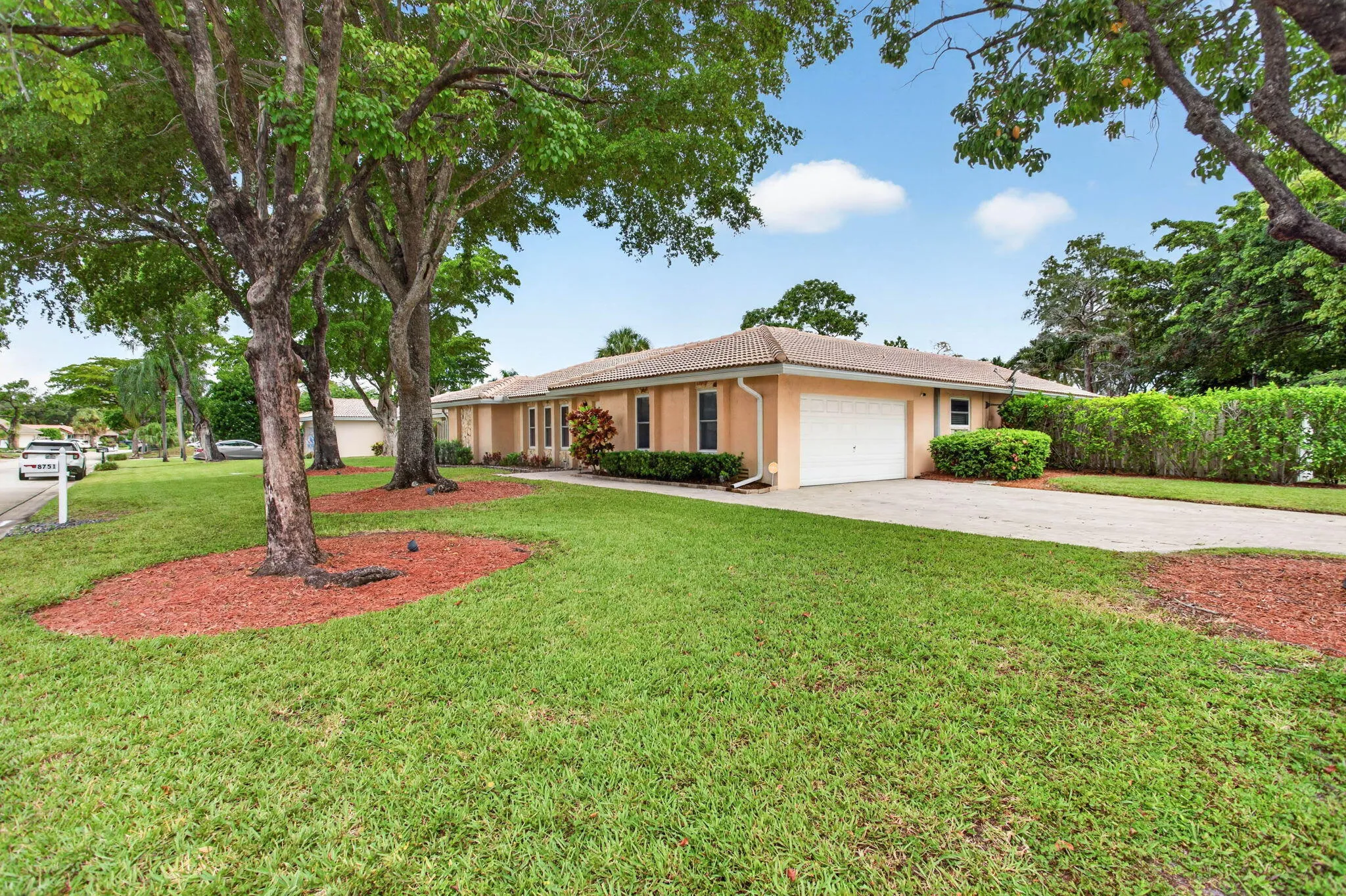 Manicured lawn with mature trees, paved driveway, stucco exterior, attached garage, and privacy hedge