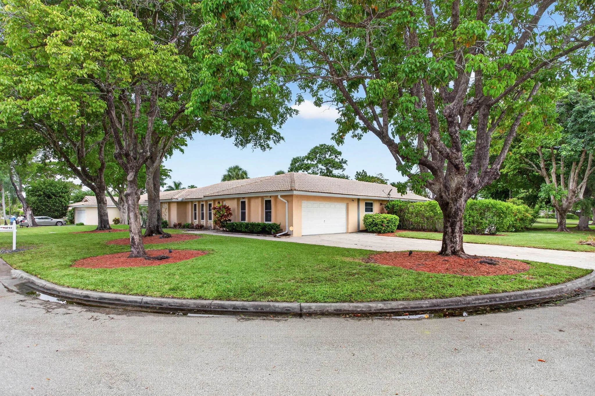 Single-story exterior with a light-colored facade, featuring multiple mature trees and a green lawn