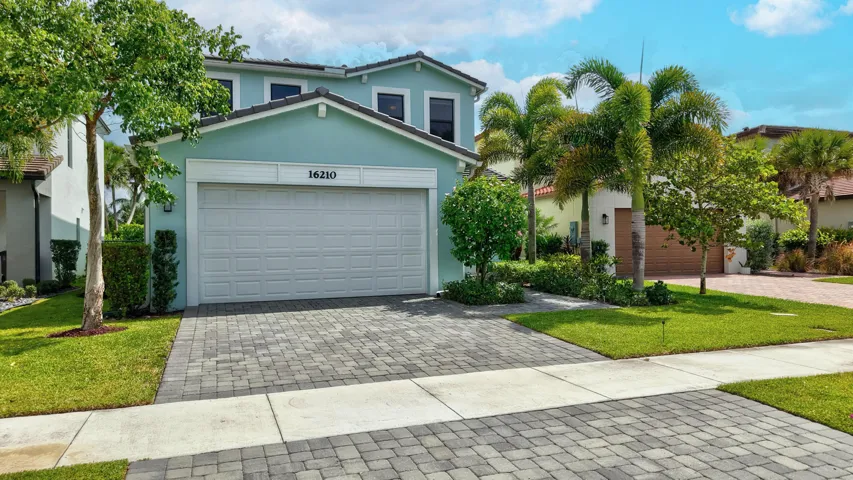 Two-story exterior with a light blue finish, featuring a paver driveway, an attached garage, and mature landscaping