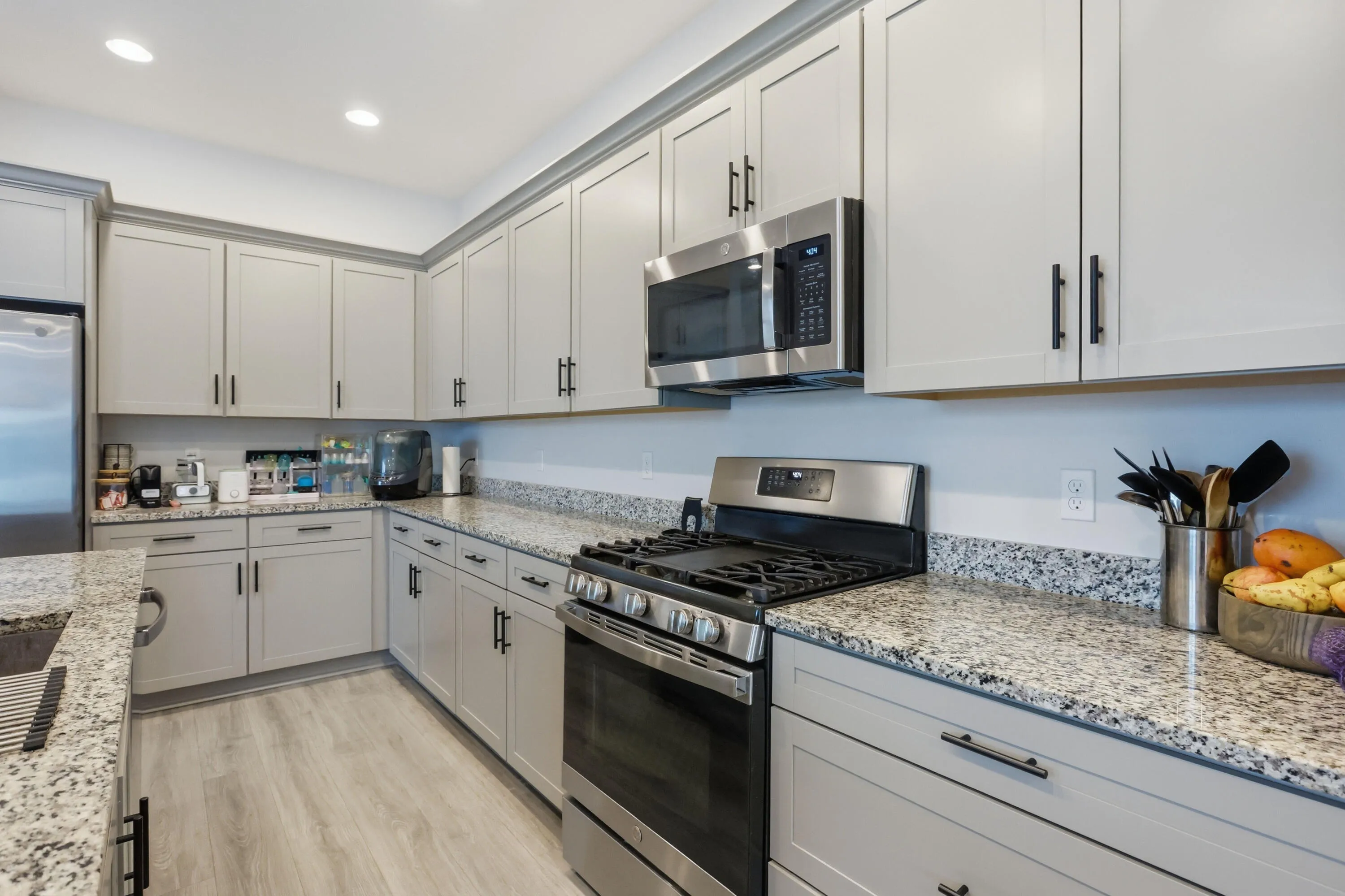 Galley kitchen featuring light-toned cabinetry with black hardware, stainless steel appliances, speckled stone countertops, and wood-finish flooring