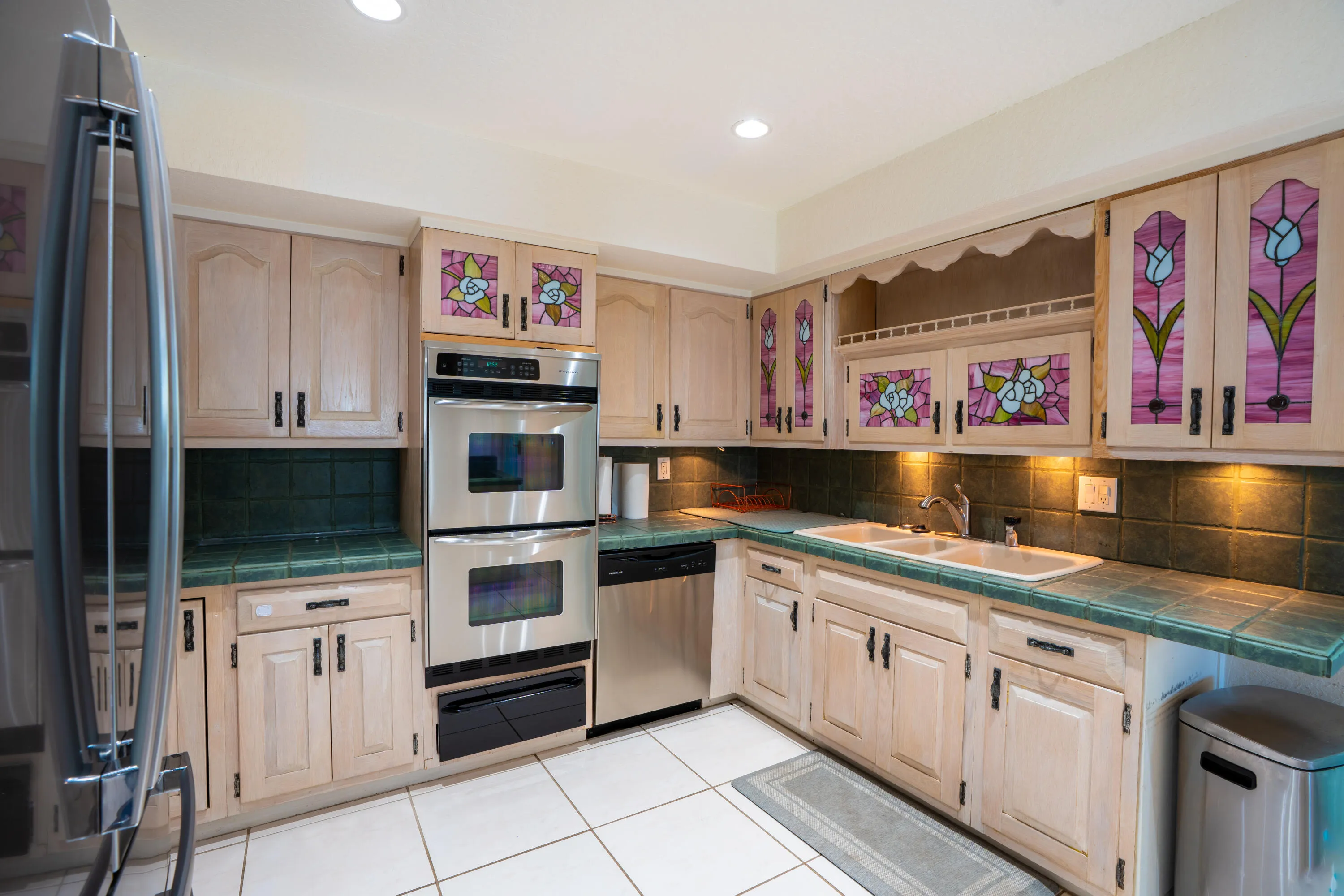 Stainless steel double oven, stainless steel refrigerator, stainless steel dishwasher, tile flooring, and custom cabinetry with stained glass inserts