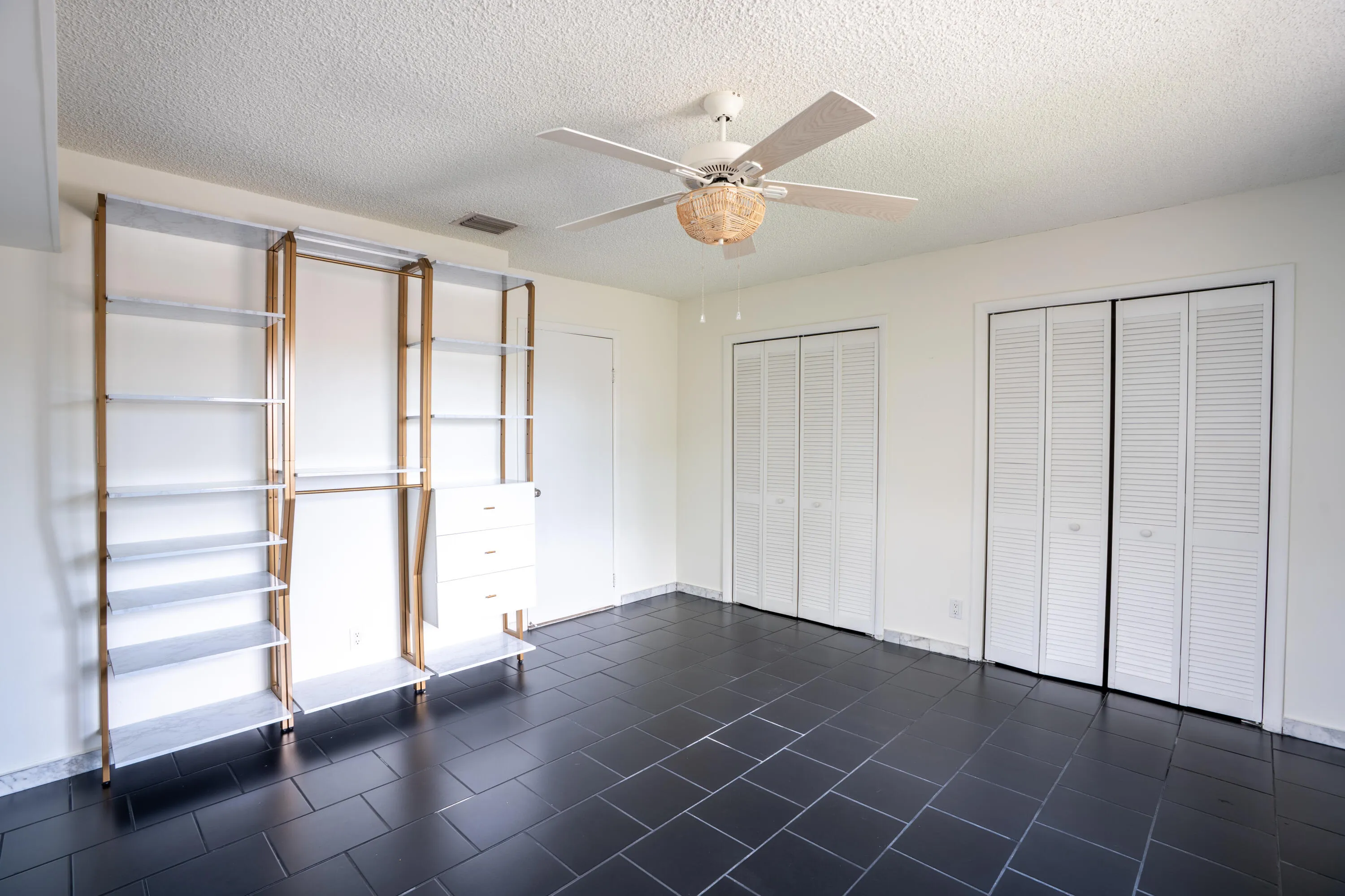 Black tile flooring, built-in shelving system with hanging rods and drawers, two louvered bifold closet doors, white ceiling fan, and white walls