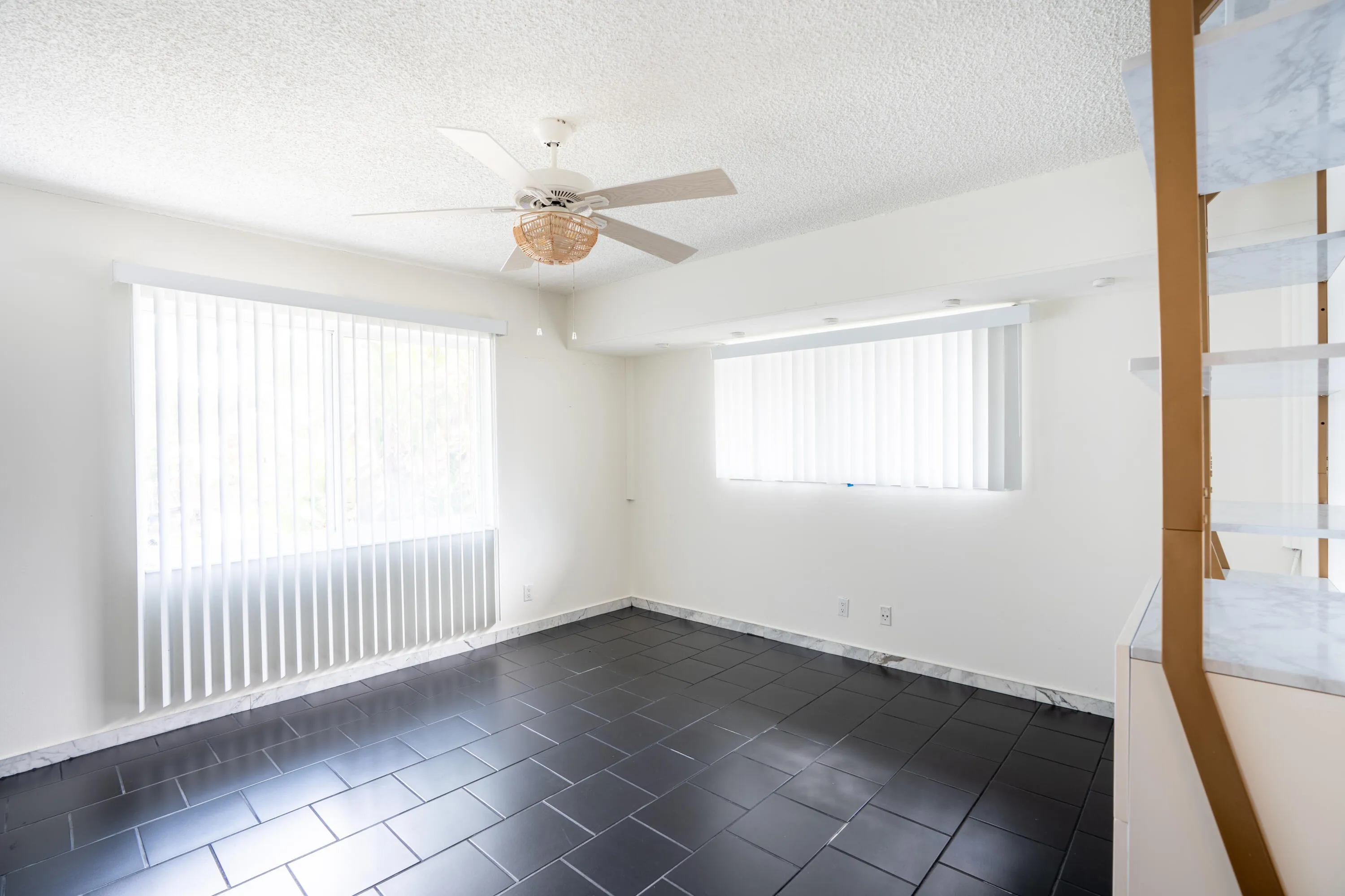Dark tile flooring, white walls, ceiling fan, multiple windows with vertical blinds, and built-in shelving