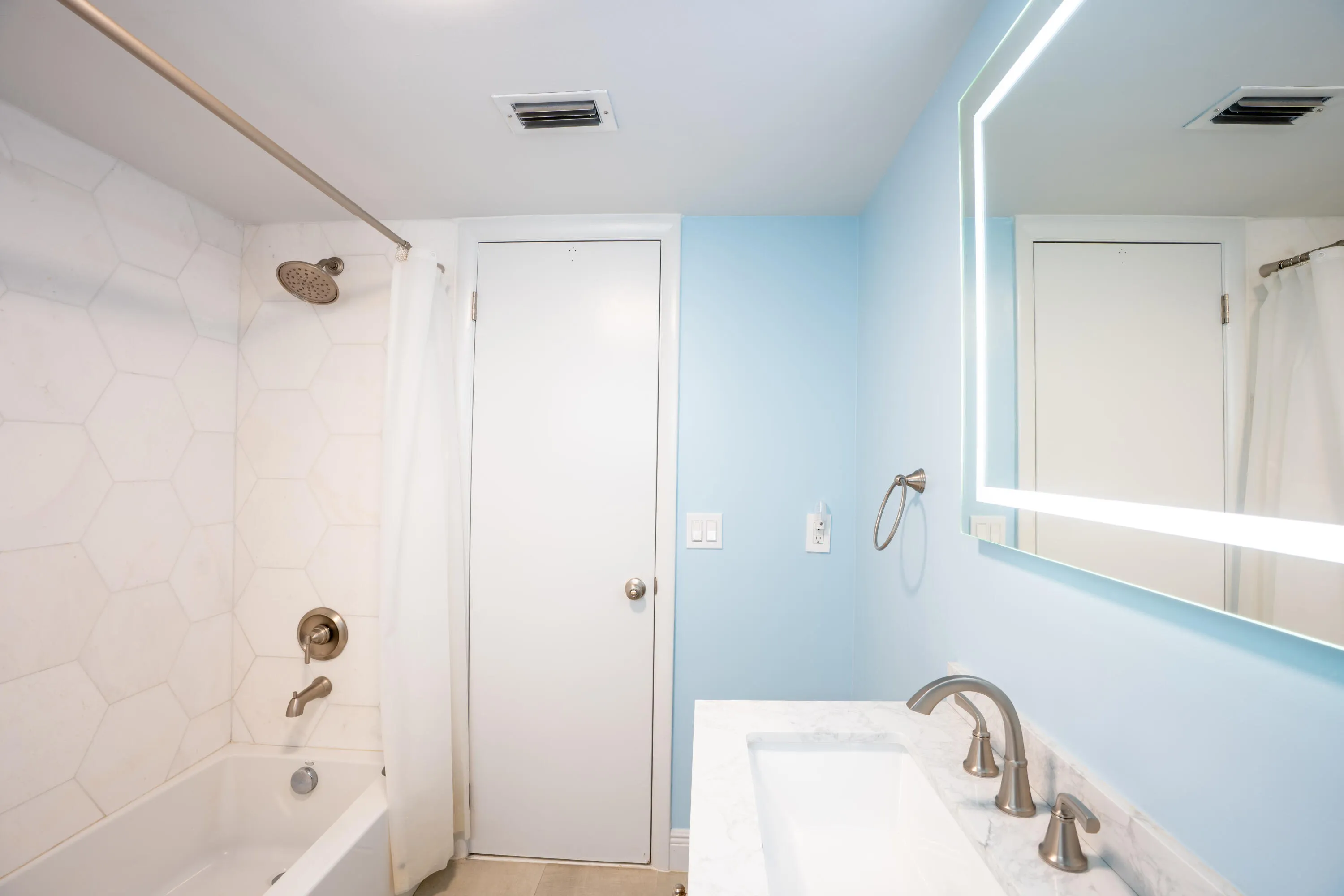 Bathtub with hexagonal tile surround, brushed nickel shower fixtures, vanity with integrated rectangular sink, marble-finish countertop, and illuminated mirror