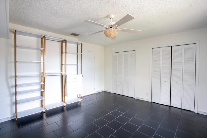 Black tile flooring, built-in shelving system with hanging rods and drawers, two louvered bifold closet doors, white ceiling fan, and white walls