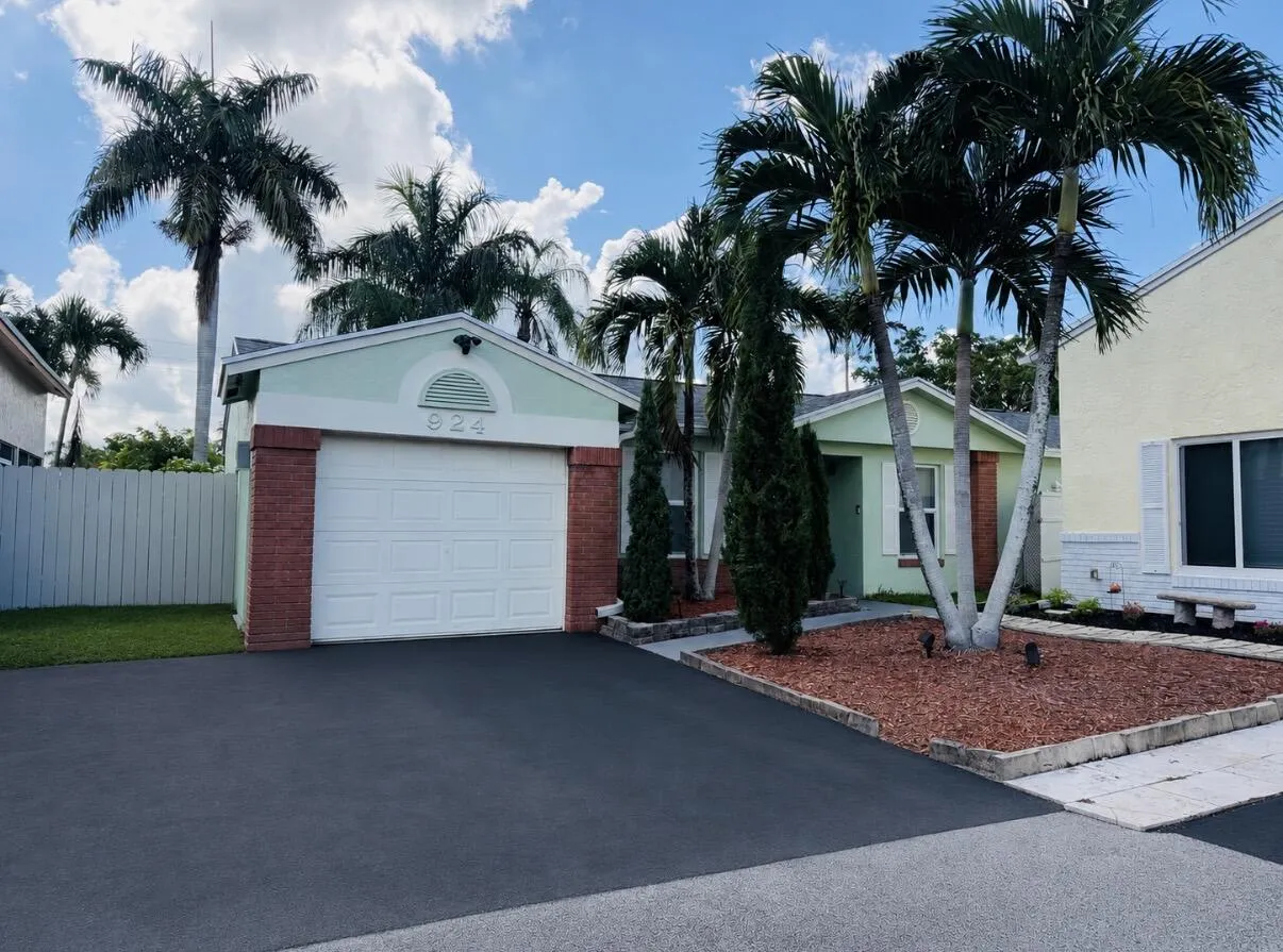 Exterior featuring a paved driveway, attached garage, brick facade accents, and mature palm trees