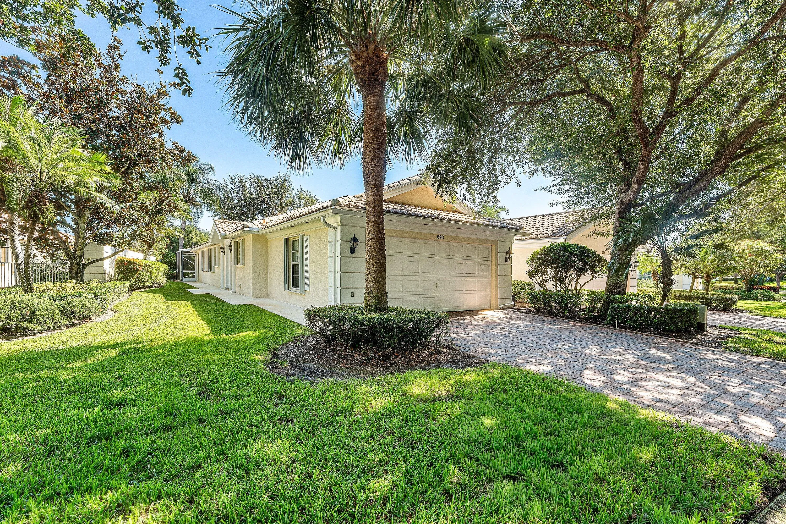 Stucco exterior with a tiled roof, featuring a paver driveway, an attached garage, and mature landscaping
