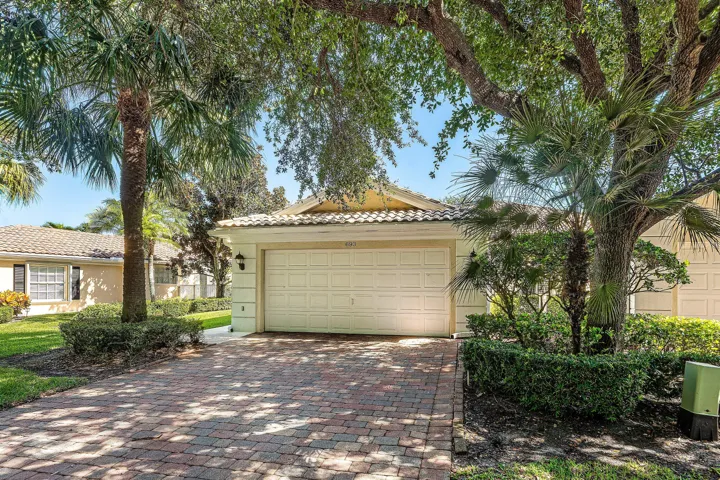 Brick paver driveway, attached two-car garage, mature landscaping, tiled roof, and exterior lighting