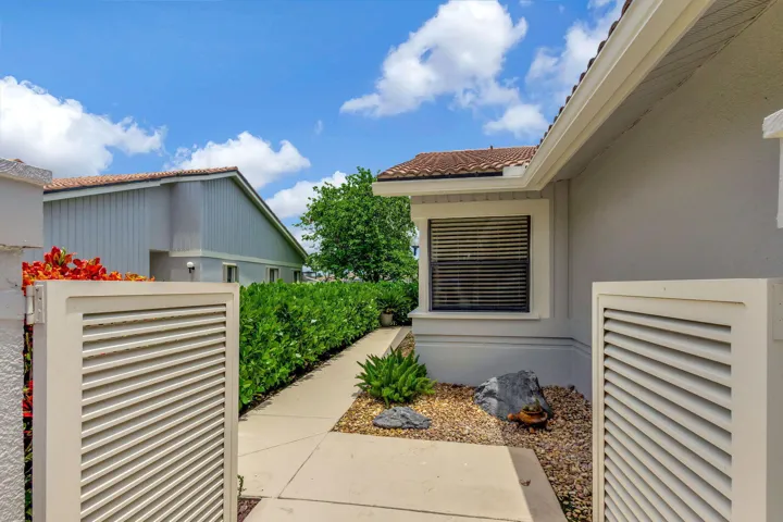 Gated entryway with a concrete pathway, privacy hedging, exterior rock landscaping, and a window with horizontal blinds