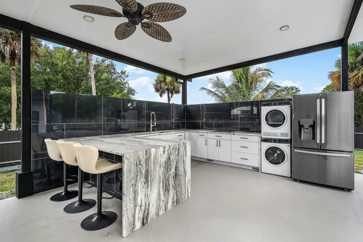 Covered outdoor kitchen featuring a large stone-finish island, white cabinetry, dark tile backsplash, a stacked washer and dryer, and a stainless steel refrigerator