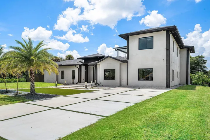 Contemporary architectural design with a multi-level structure, a concrete driveway featuring grass inlays, a large front lawn, and a prominent palm tree