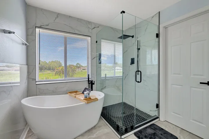 Freestanding soaking tub with a large window, glass-enclosed shower with black hexagonal tile, and black fixtures, complemented by marble-patterned wall and floor tiling