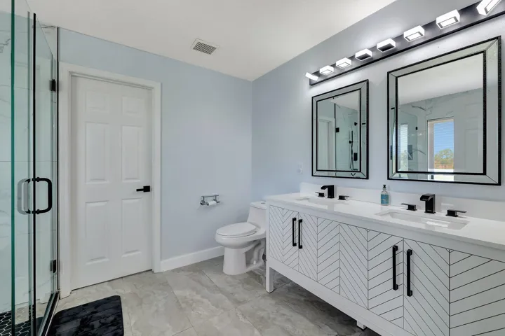 Dual vanity with white countertops and matte black fixtures, framed mirrors, and a glass-enclosed shower with black hexagonal tile flooring