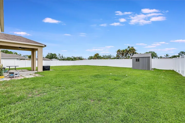 Expansive grassy backyard featuring a white privacy fence, a concrete patio, and a covered outdoor area