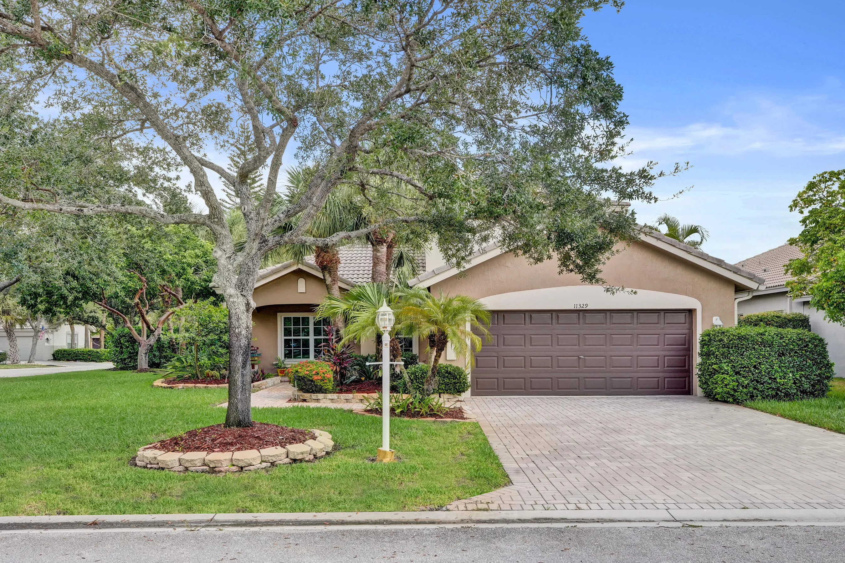 Welcoming curb appeal featuring a mature shade tree with circular mulch bed, manicured lawn, and professional hardscape paver driveway