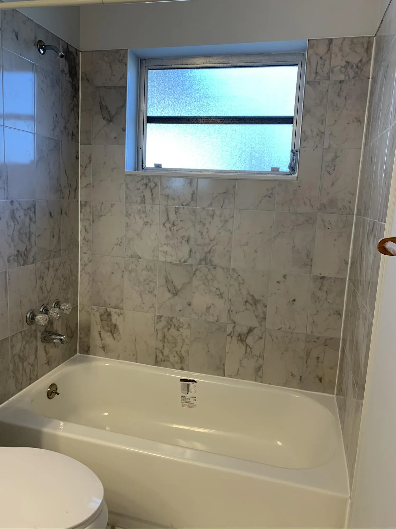 Bathtub with a tiled surround, frosted glass window, and wall-mounted shower fixtures