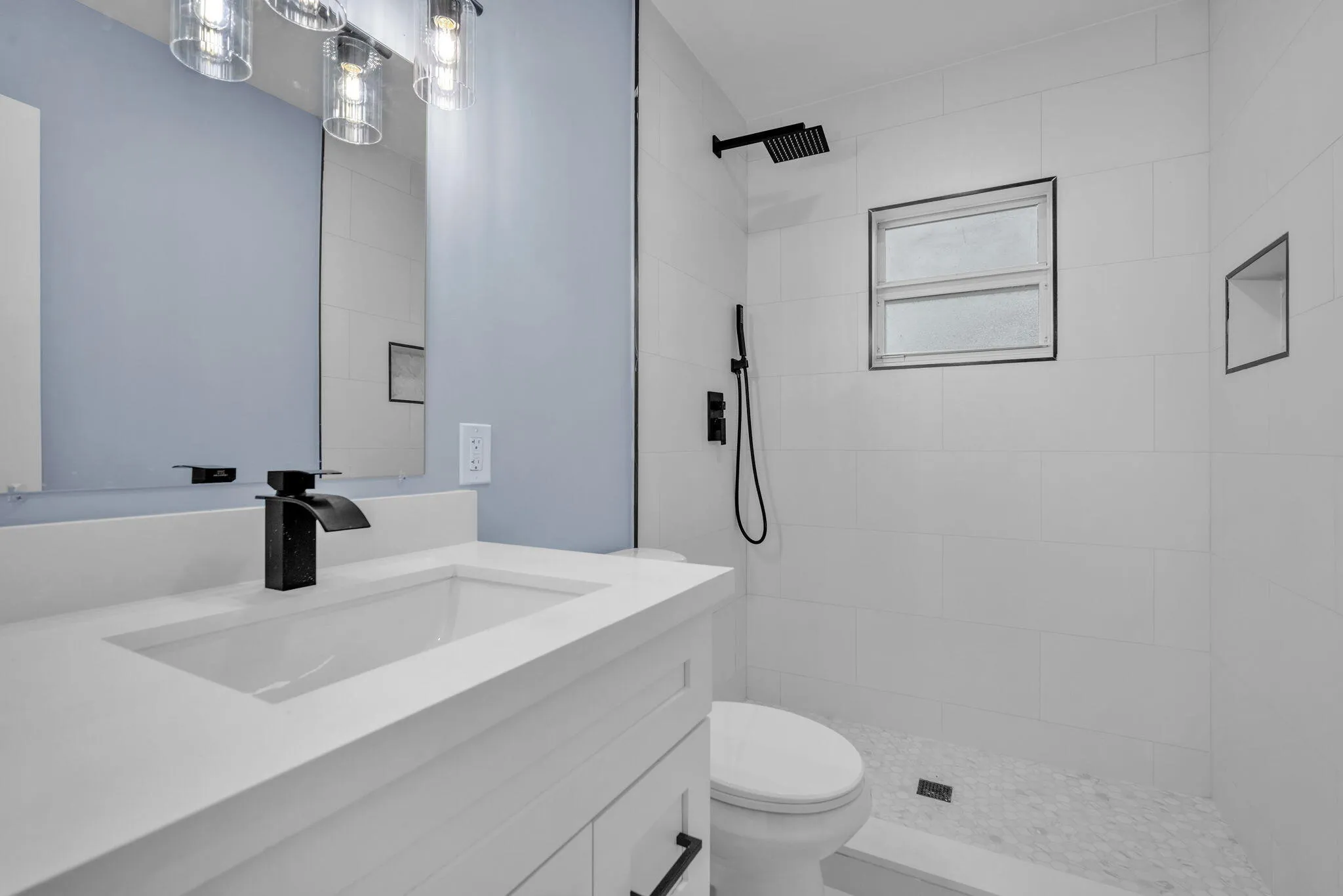 Vanity with integrated sink and matte black faucet, wall-mounted mirror, three-light vanity fixture, white tiled shower with rain shower head, and built-in shower niche