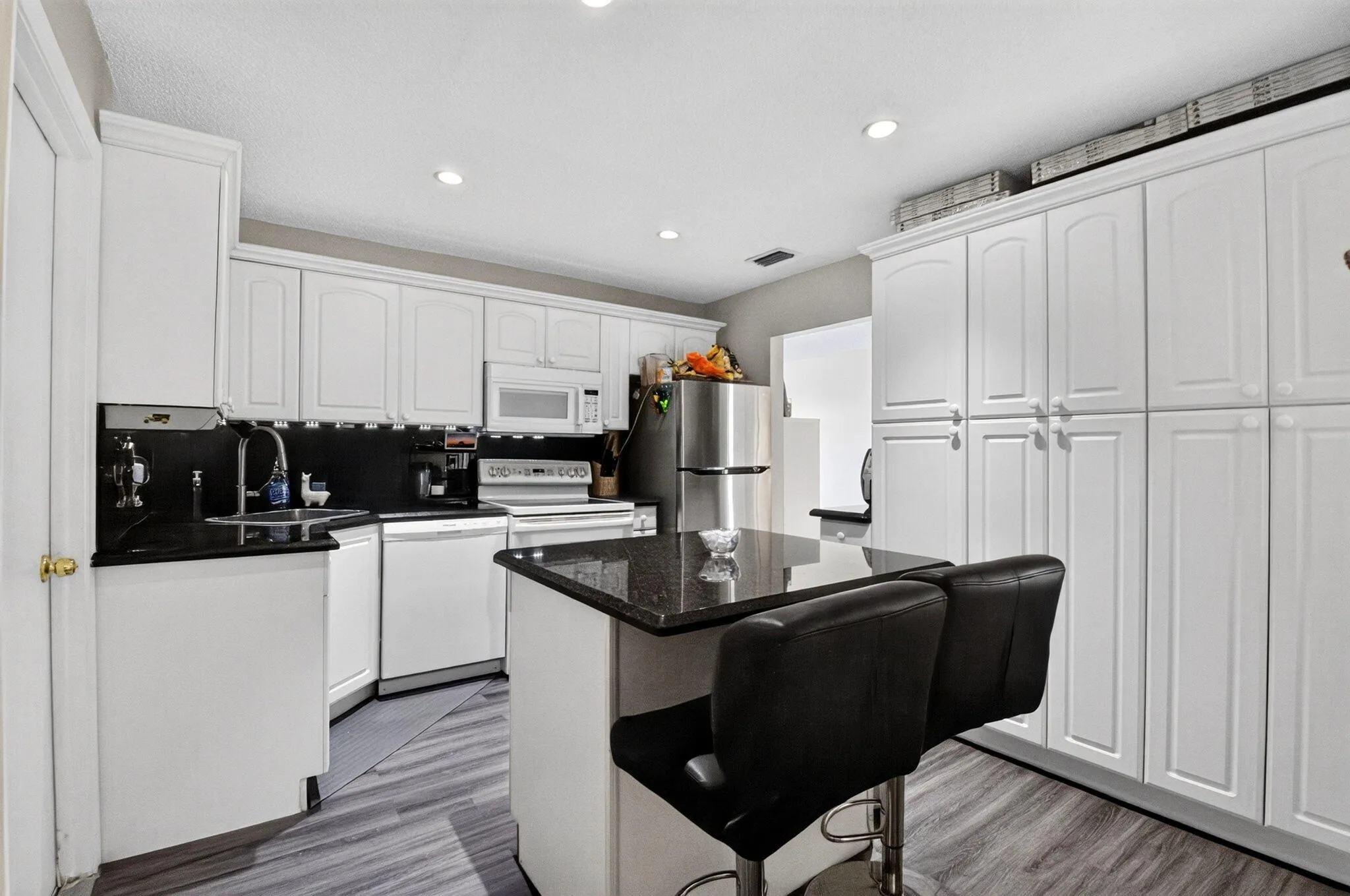 Modern kitchen featuring white shaker-style cabinetry, stainless steel appliances, black granite countertops, a center island with seating, and wood-finish flooring throughout the open-concept space
