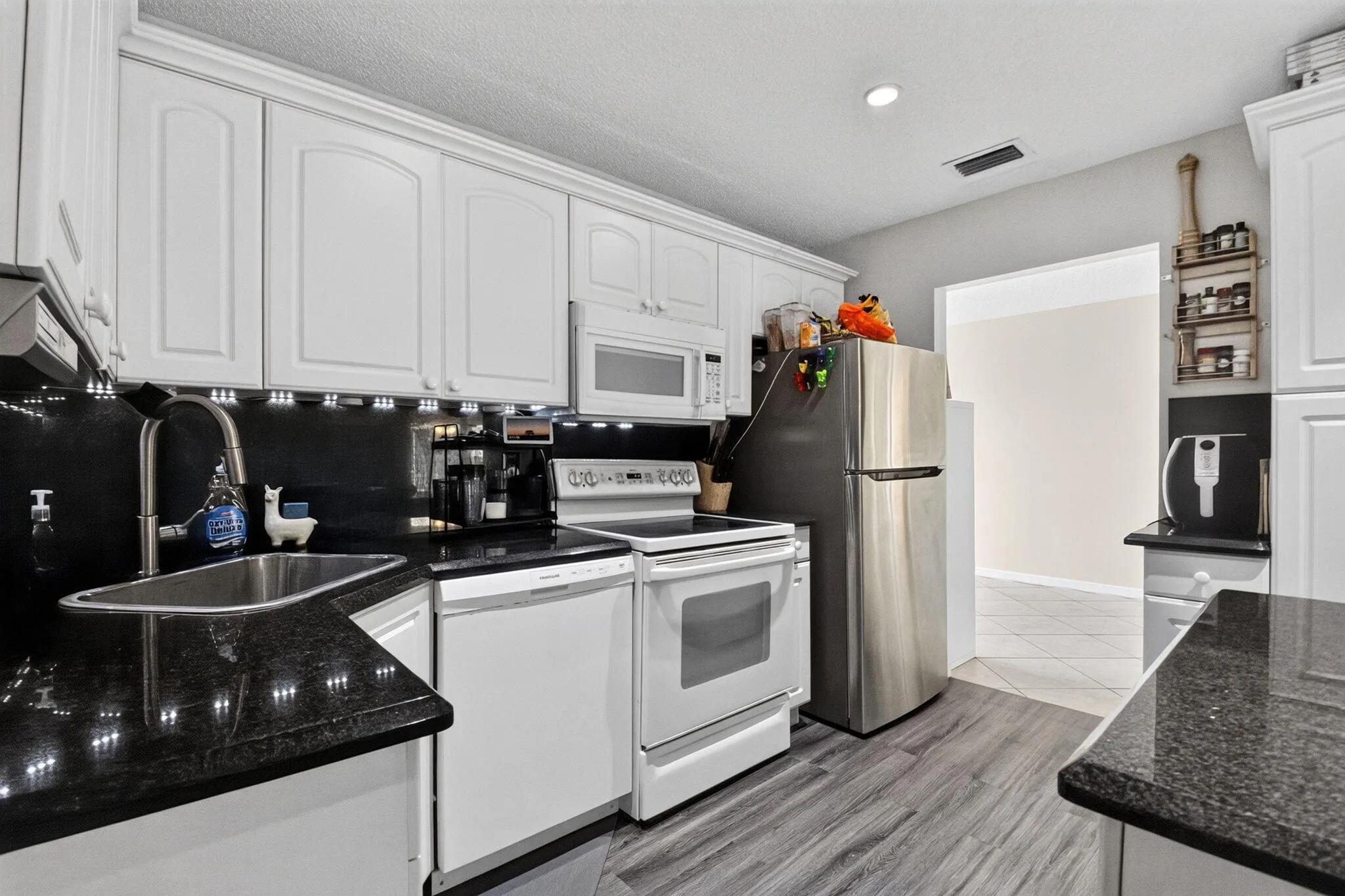 White cabinetry with crown molding, dark speckled countertops, stainless steel sink, under-cabinet lighting, and wood-finish flooring