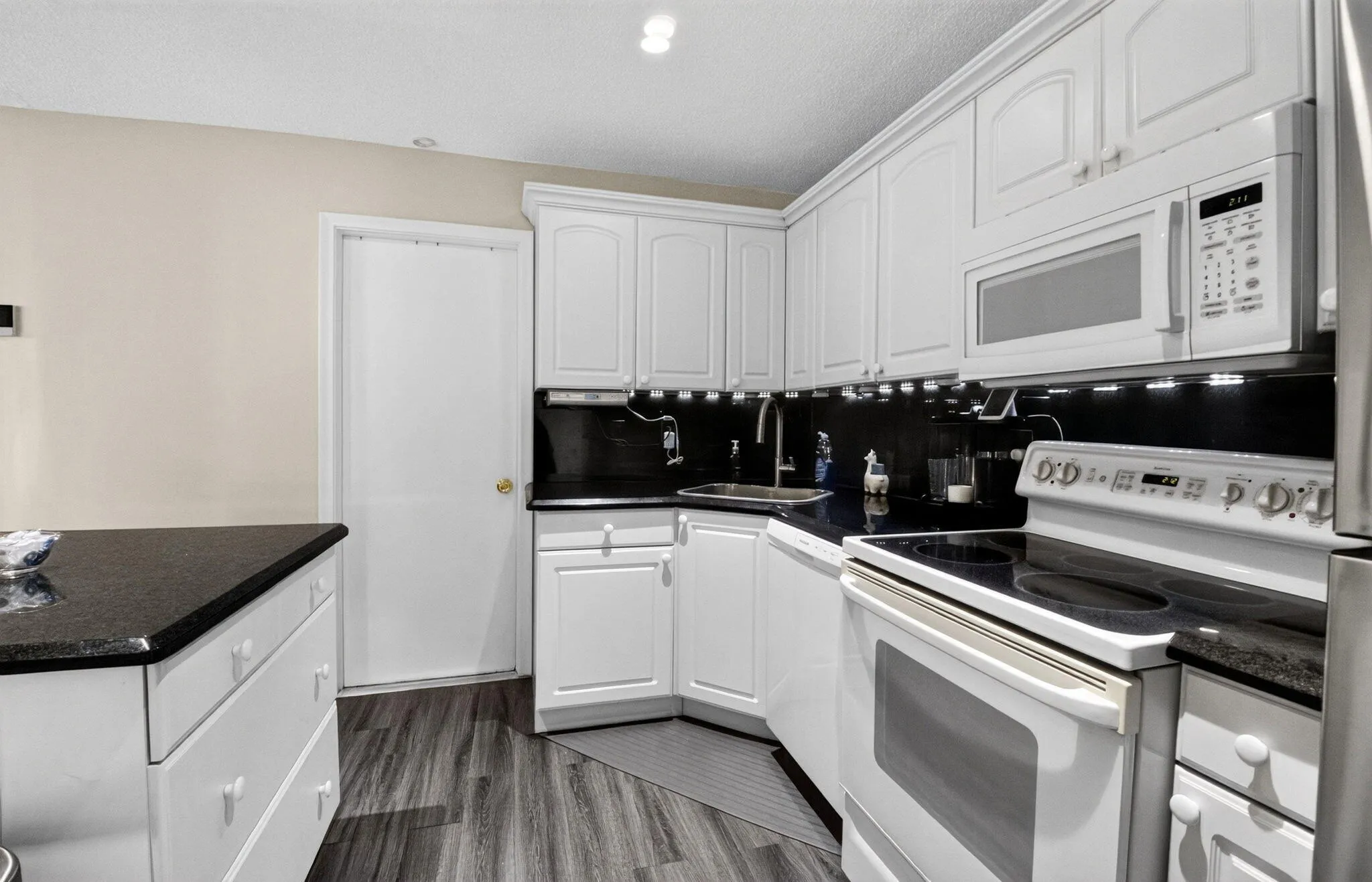 White cabinetry, dark stone countertops, wood-finish flooring, stainless steel sink, and white appliances