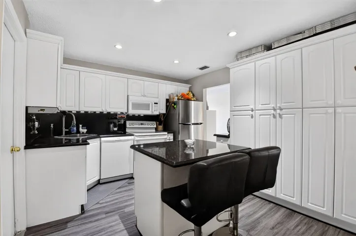 Modern kitchen featuring white shaker-style cabinetry, stainless steel appliances, black granite countertops, a center island with seating, and wood-finish flooring throughout the open-concept space