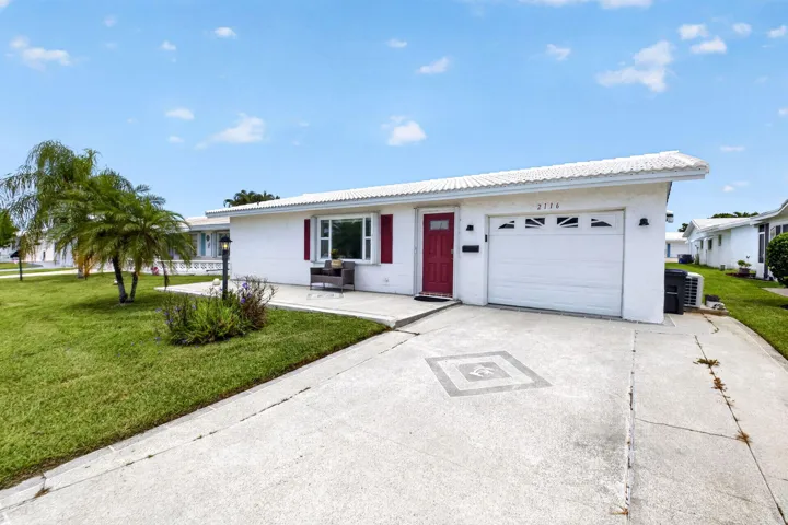 White stucco exterior with a red entry door, attached single-car garage, concrete driveway with decorative inlay, and a front patio