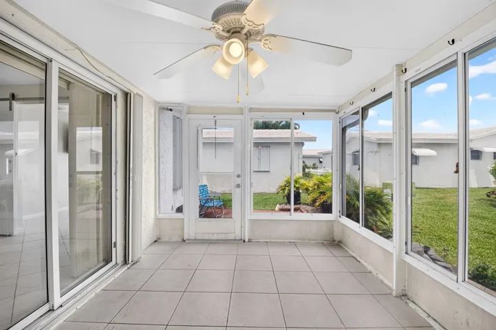 Bright sunroom showcasing expansive windows framing manicured lawn vistas, neutral tile flooring, and a ceiling fan with integrated lighting
