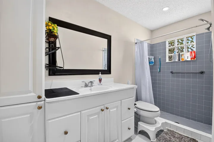 White vanity with integrated sink, rectangular mirror, full-height storage cabinetry, tiled shower with grab bar, and glass block window