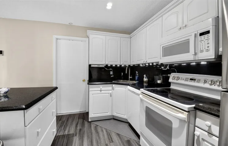 White cabinetry, dark stone countertops, wood-finish flooring, stainless steel sink, and white appliances
