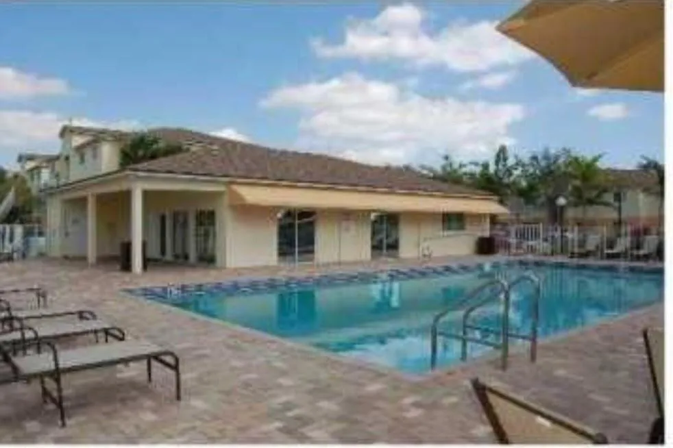 Resort-style swimming pool with crystal-blue water, expansive paved deck with lounge seating areas, mature tropical landscaping with palm trees, and a two-story clubhouse featuring covered portico and large windows overlooking the amenity