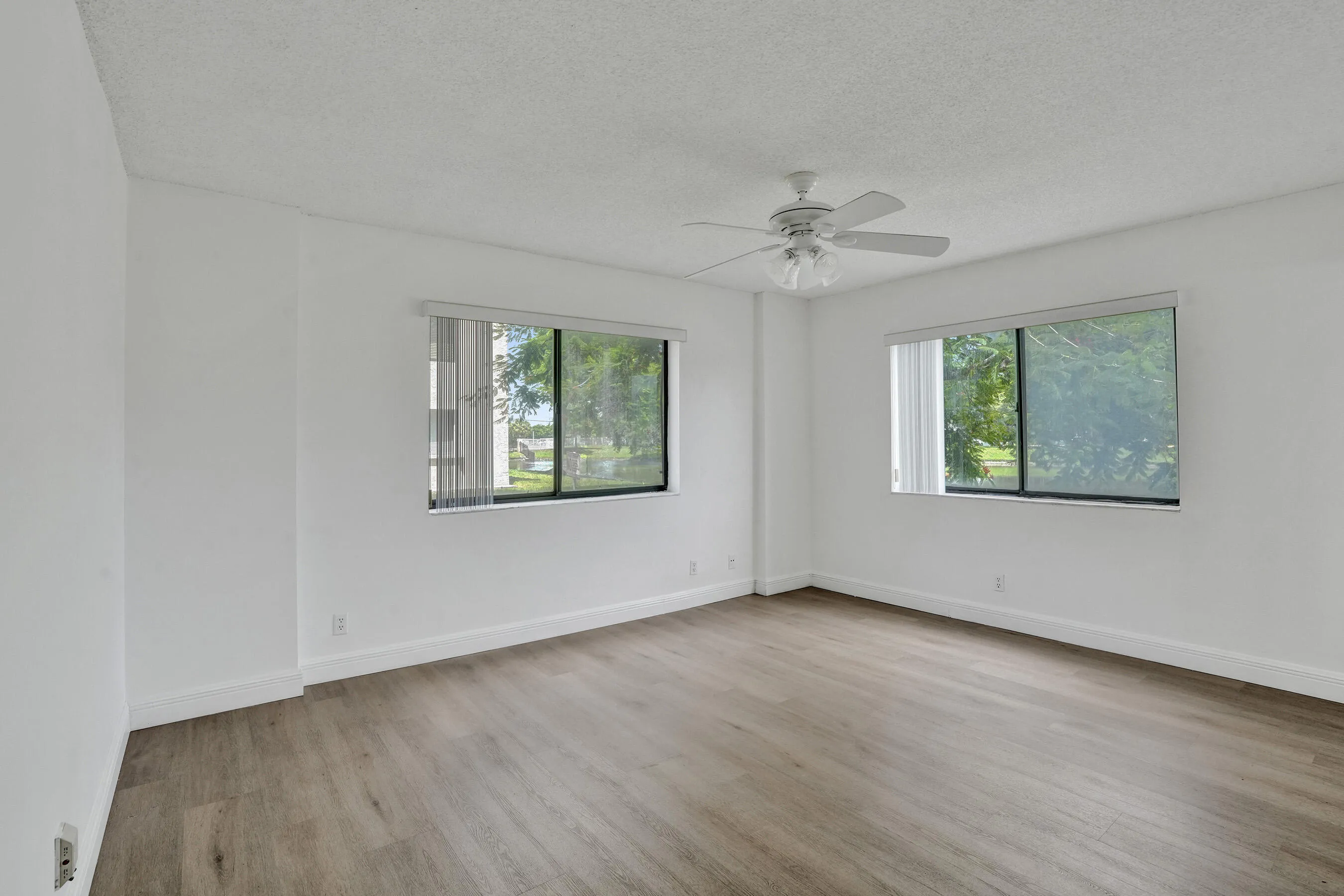 Bright, open living space featuring gleaming wood-finish flooring, dual sliding glass windows with natural light exposure, and a ceiling fan with brushed metal finish