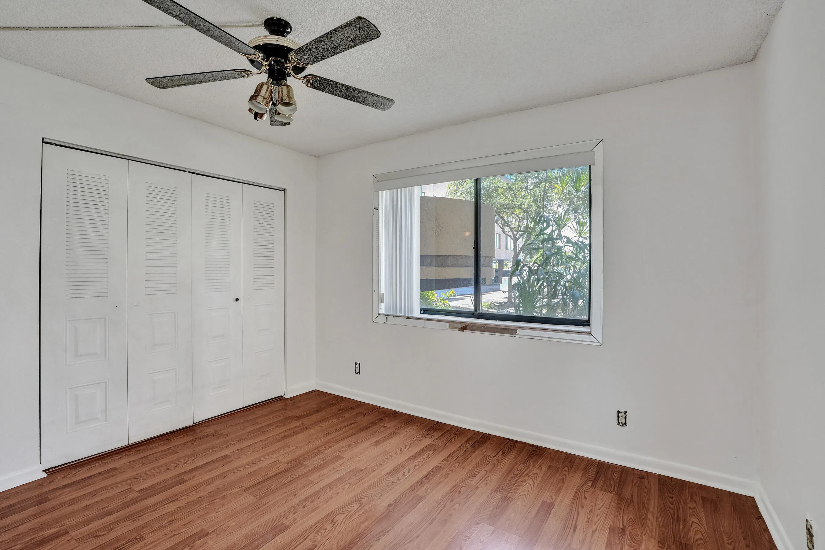Wood-finish flooring, louvered bi-fold closet doors, ceiling fan with integrated lighting, and a large window