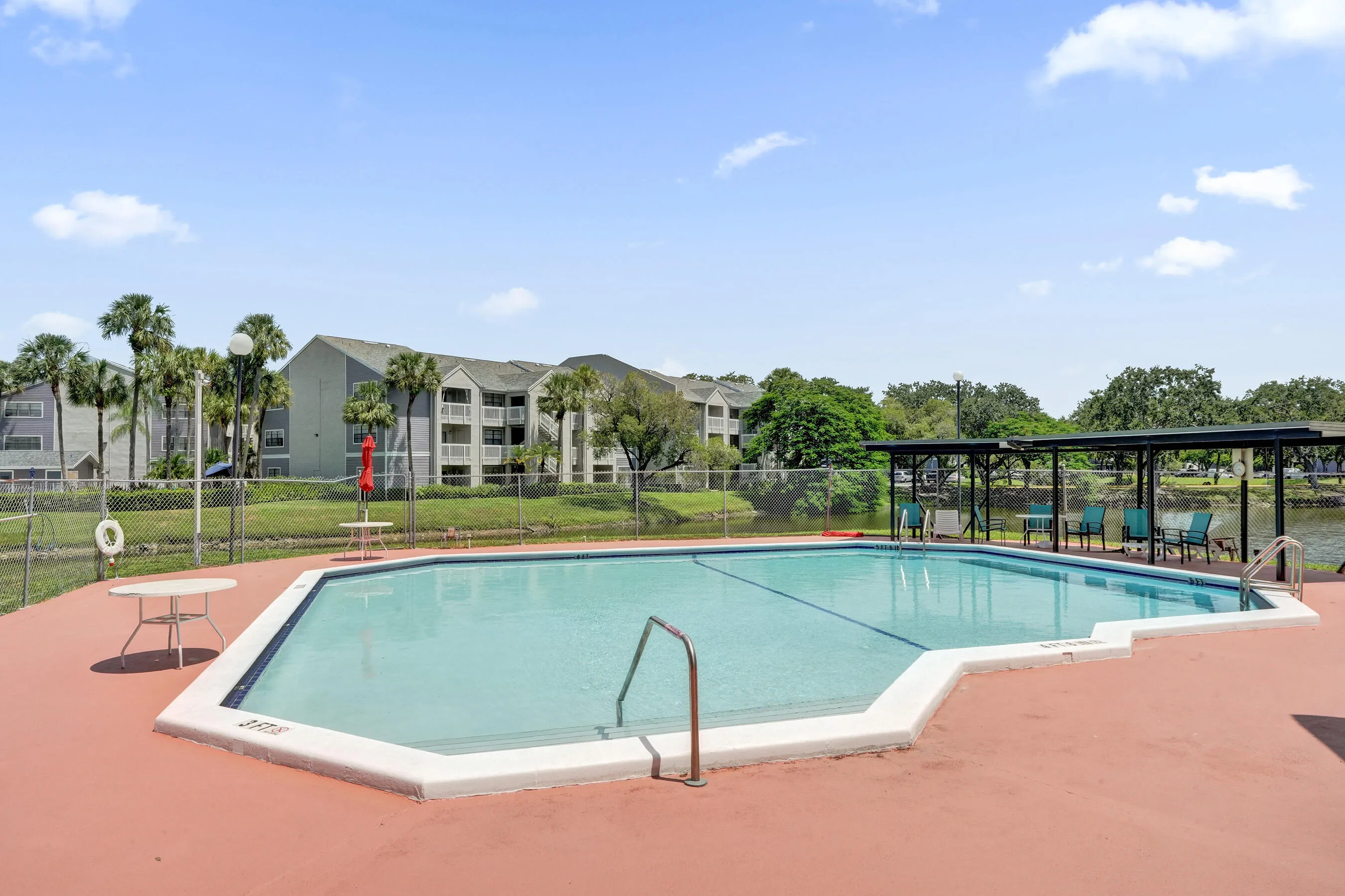 Hexagonal swimming pool with a surrounding red-toned hardscape, adjacent covered seating area, and a metal fence enclosure