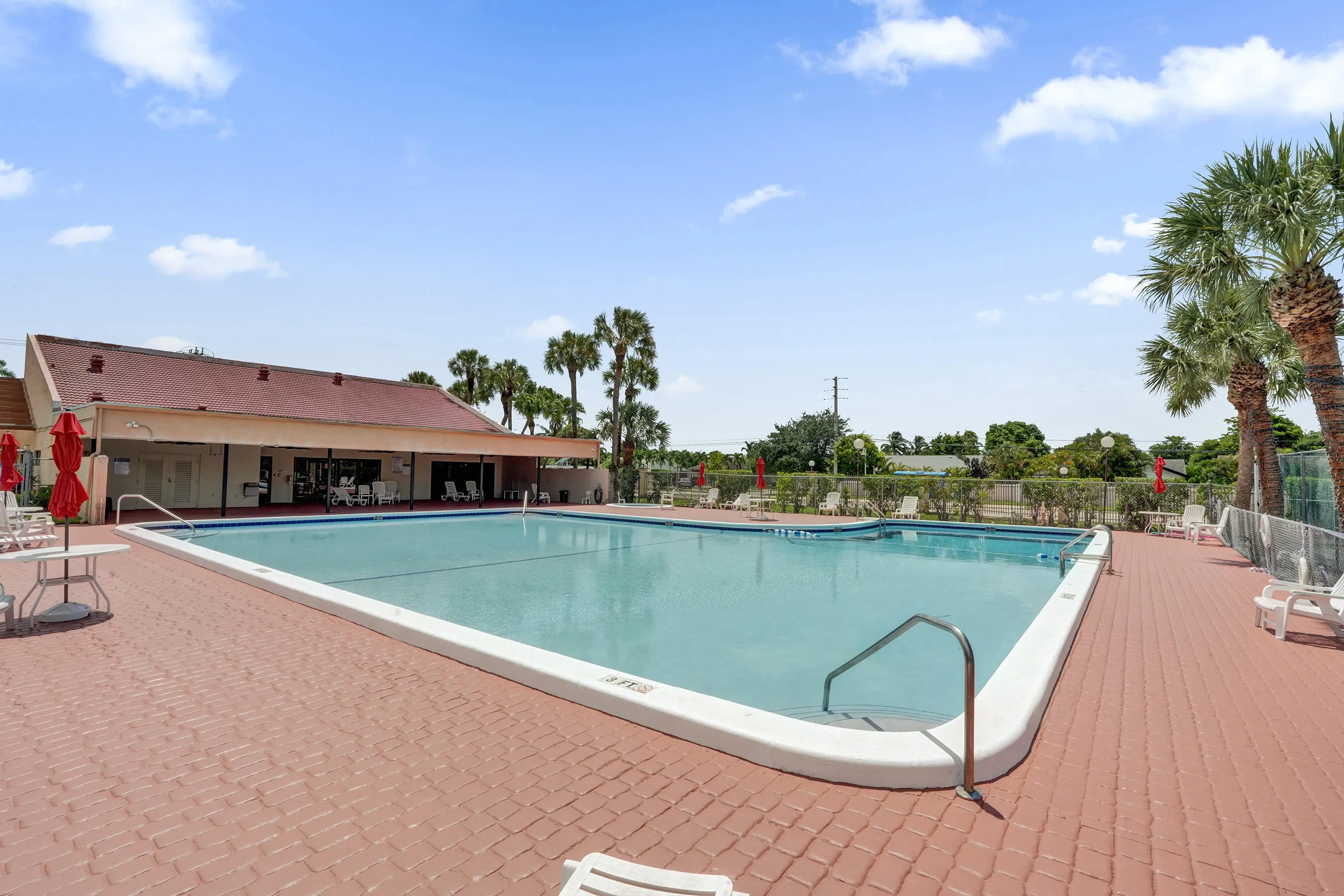Expansive outdoor pool with a red brick paver deck, surrounded by mature palm trees, and a clubhouse building