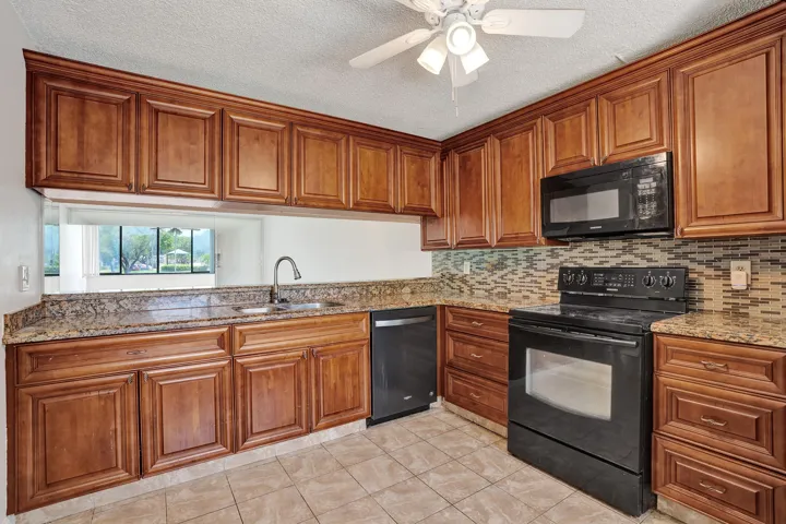 Wood-finish cabinetry, stone countertops, integrated sink with gooseneck faucet, full suite of black appliances, and tile backsplash