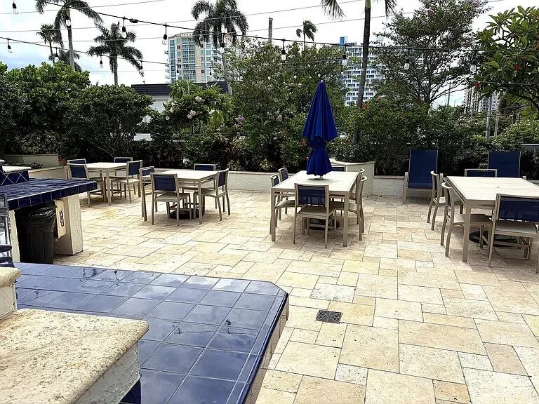 Expansive patio area featuring light-toned paver flooring, lush perimeter landscaping, and string lighting overhead