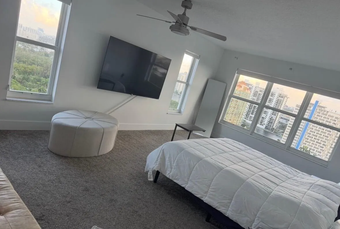 Wall-mounted television with visible wiring conduit, neutral-toned carpet, white walls, and multiple windows providing cityscape and greenery views
