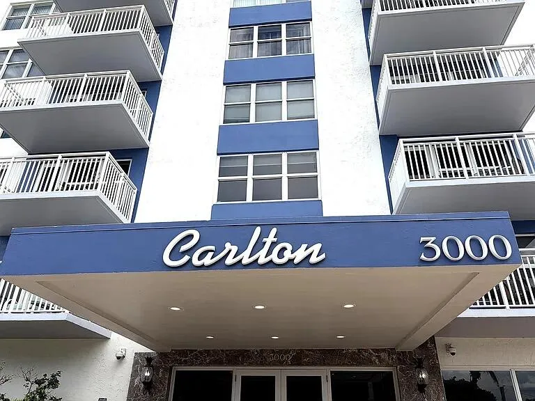 Building exterior with multiple balconies featuring white railings, blue and white facade, and a prominent entrance canopy