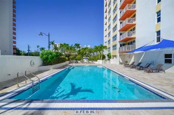 Rectangular swimming pool with integrated steps, surrounded by a light-colored paver deck, adjacent to a multi-story building featuring private balconies, with a blue fabric sunshade
