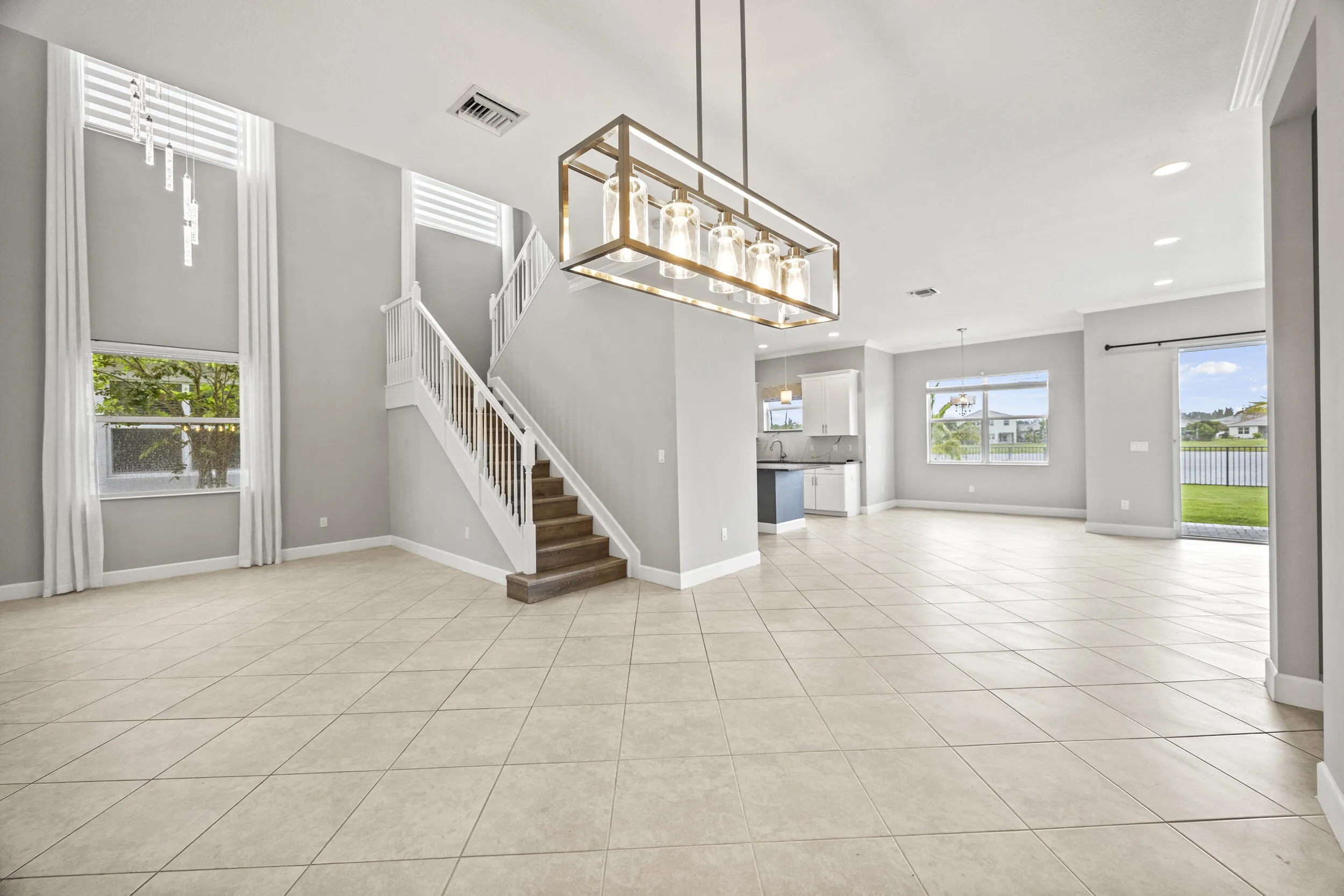 Expansive open-concept living area featuring large format tile flooring, a prominent staircase with wood-finish treads, and a modern linear chandelier
