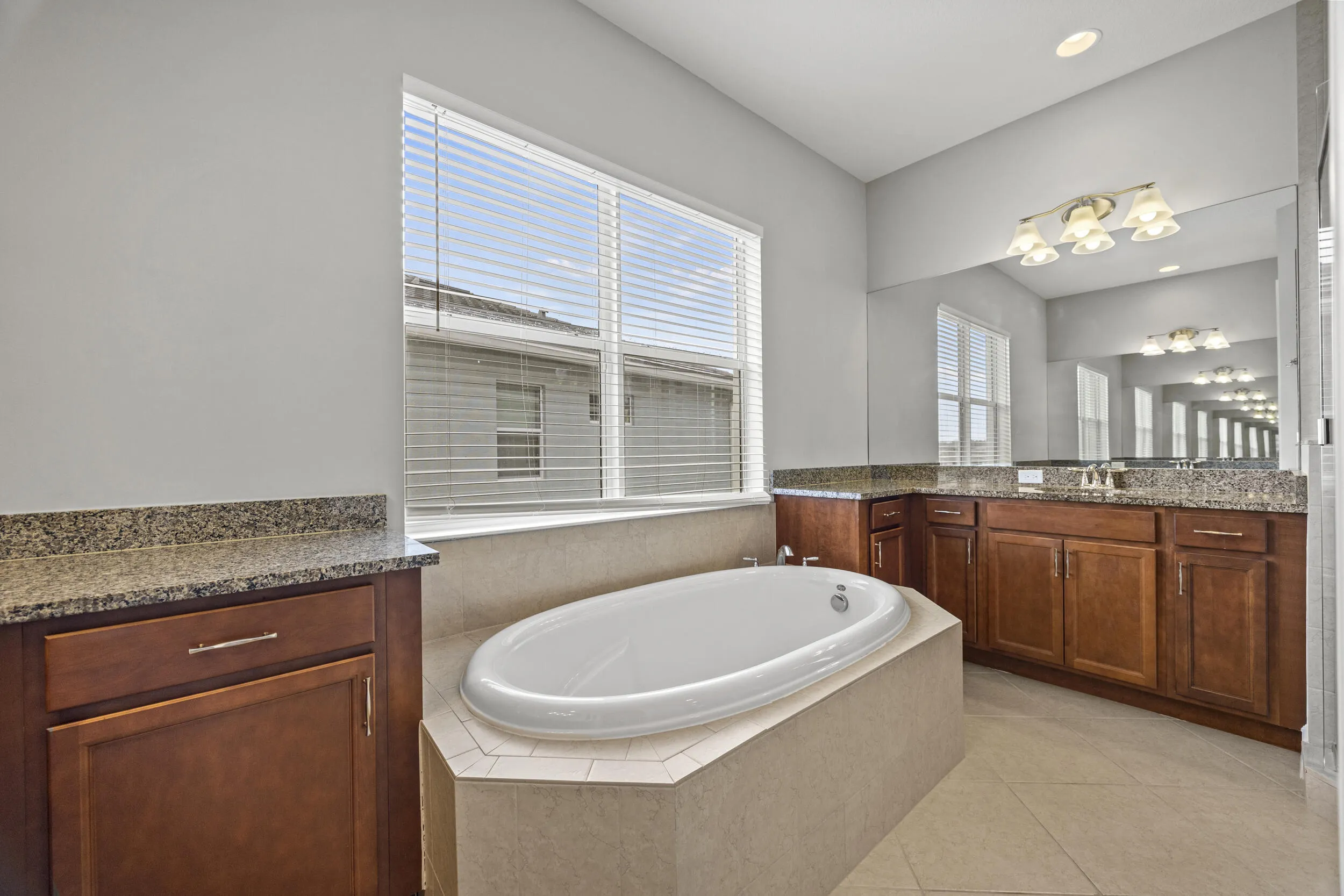 Soaking tub set within a tiled surround, dual wood-finish vanities with stone countertops, and a large mirror