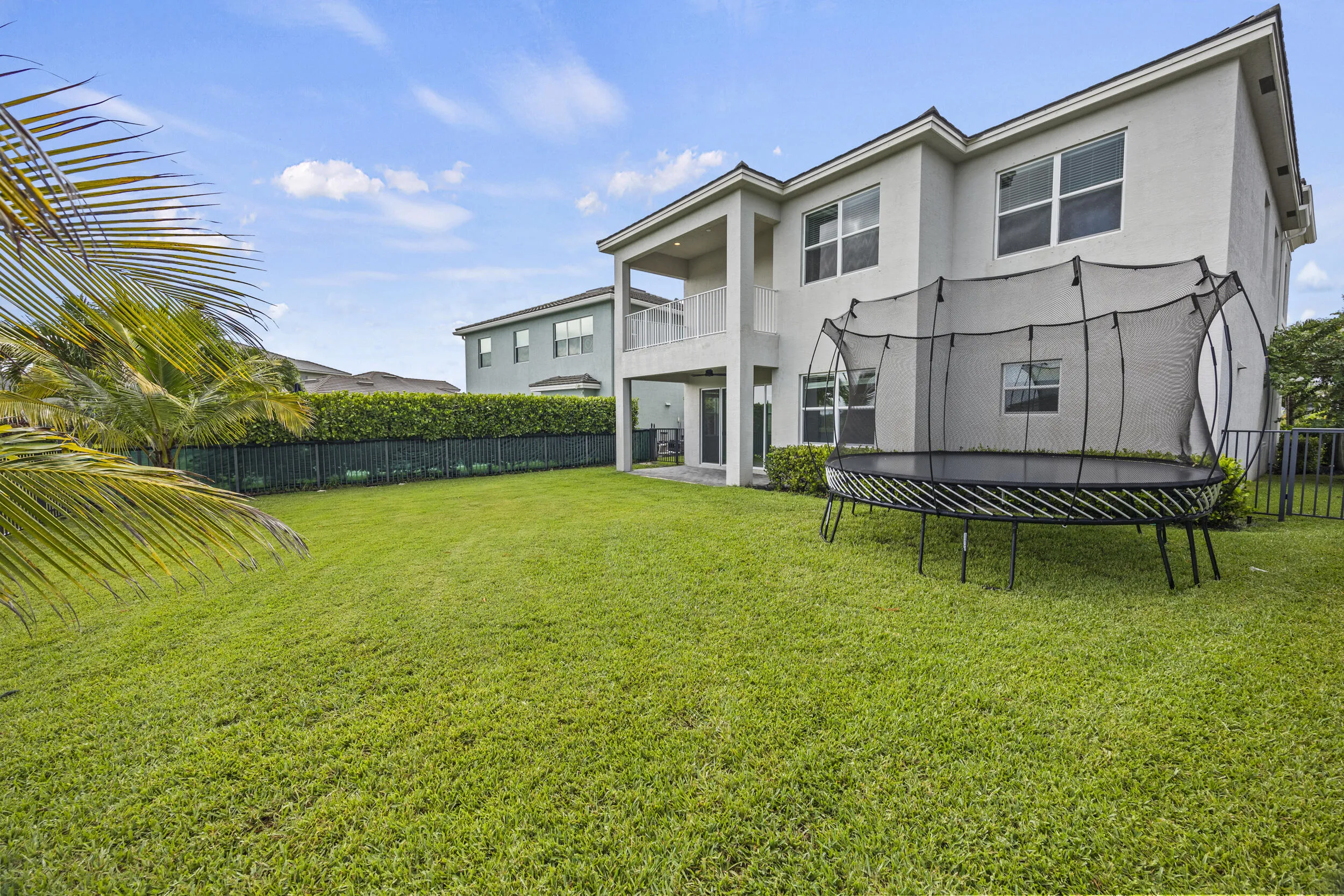 Expansive grassy yard with a two-story stucco exterior, upper-level balcony, and a dark metal fence with privacy hedges