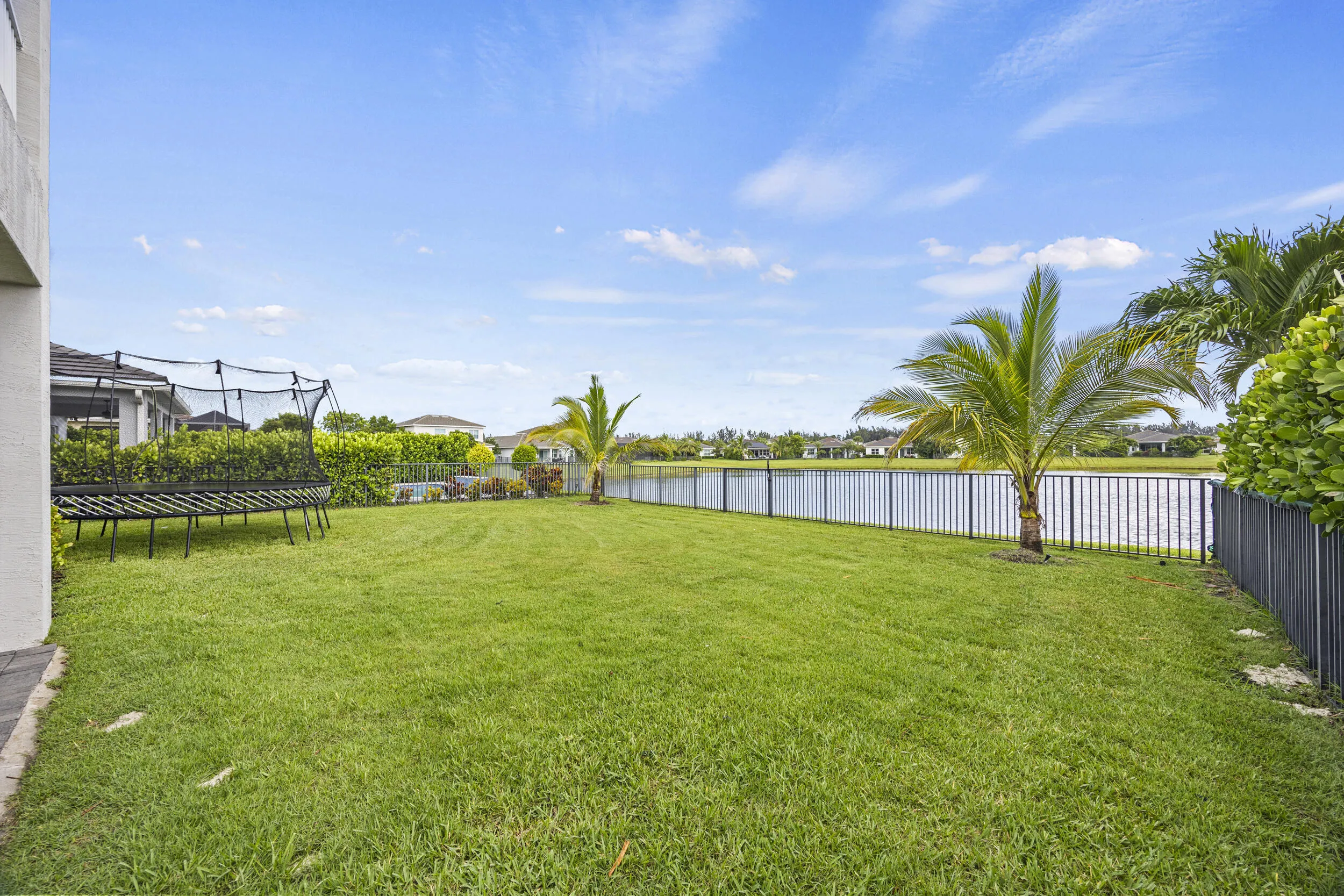 Expansive green lawn with a black metal fence bordering a water feature, featuring mature palm trees and landscaped shrubbery