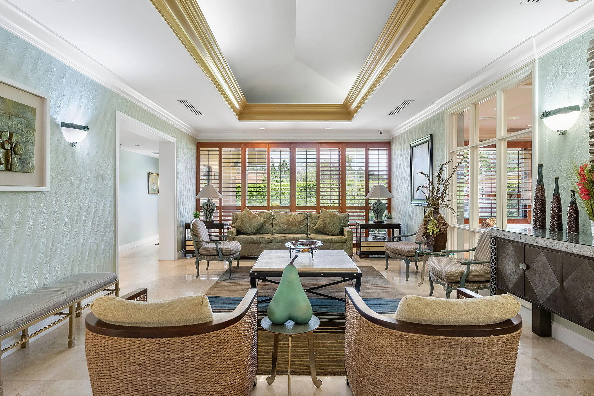 Sophisticated living room showcasing coffered ceiling with brass trim, light wood-finish shutters spanning expansive windows, neutral marble-finish flooring, woven natural fiber seating, and a geometric area rug anchoring the space