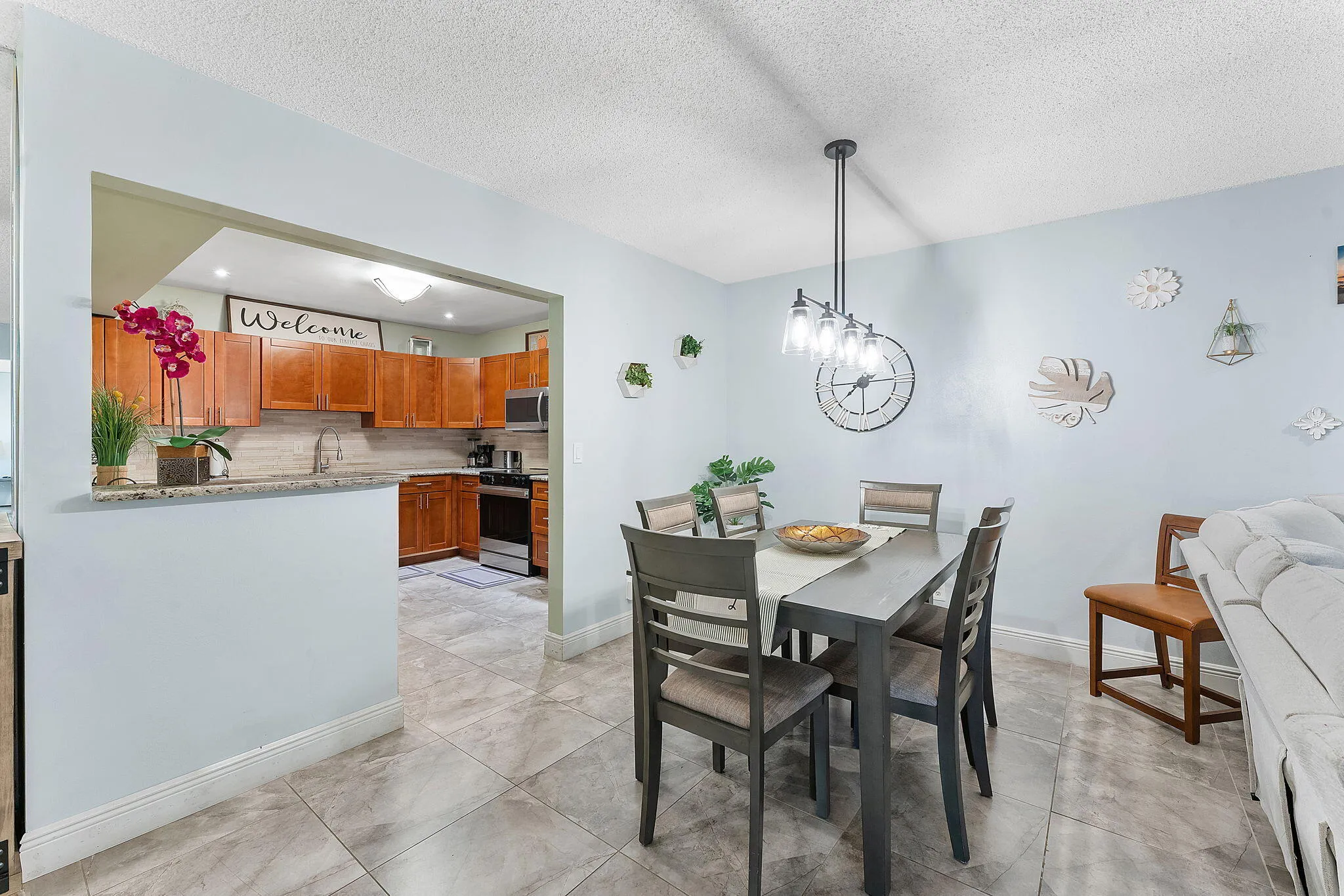 Open-concept dining and kitchen area featuring a wood-finish dining table with ladder-back chairs, stainless steel appliances, warm wood cabinetry, tile flooring, and a pass-through window to the kitchen