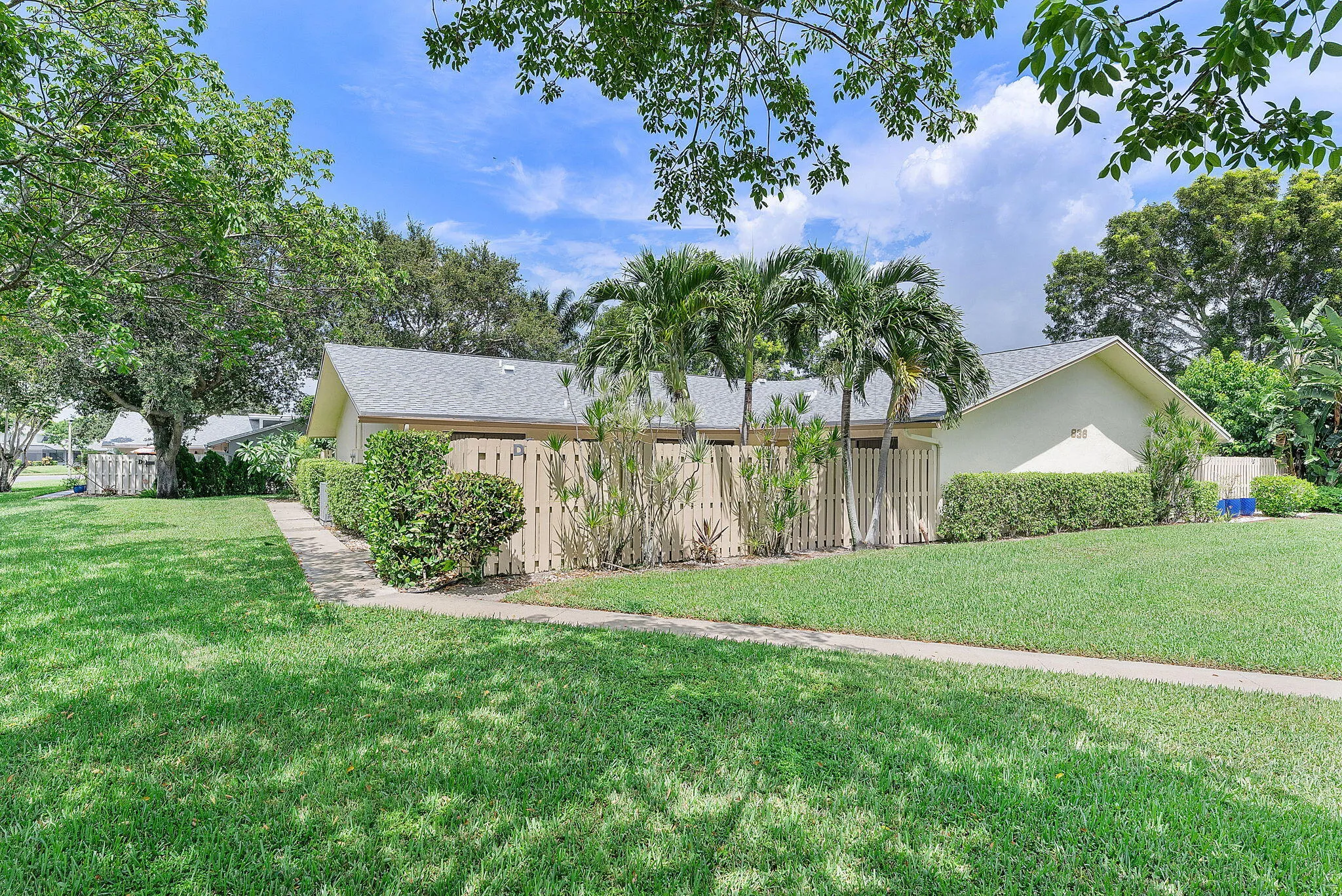 Sprawling grounds featuring mature palm trees and lush landscaping, anchored by a single-story residence with a pitched roof and wood-finish fencing