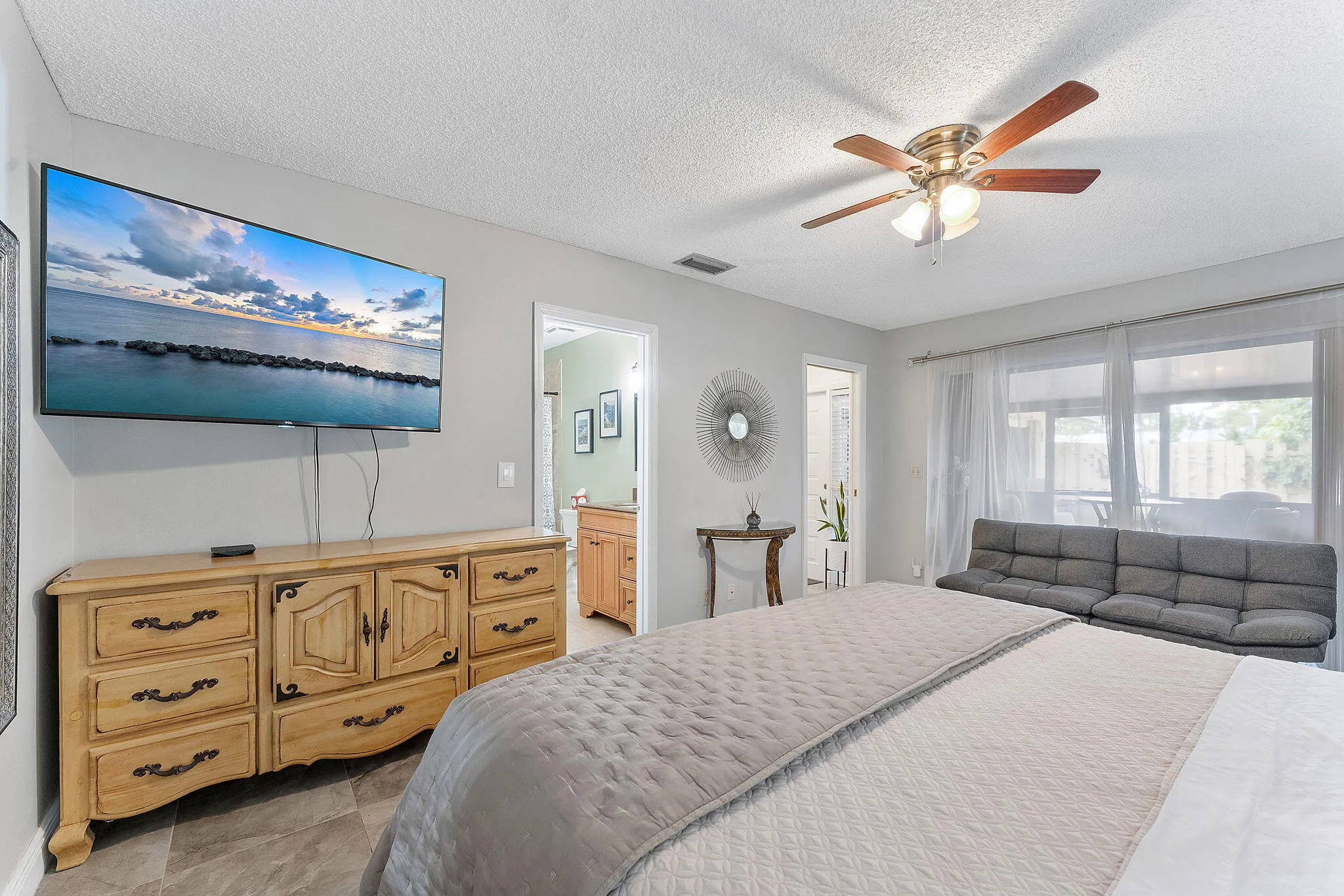 Spacious primary bedroom featuring abundant natural light from multiple windows, neutral wall finishes, wood-finish dresser with decorative hardware, ceiling fan with integrated lighting, and plush bedding