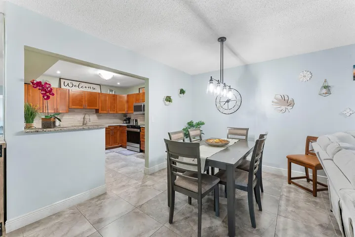 Open-concept dining and kitchen area featuring a wood-finish dining table with ladder-back chairs, stainless steel appliances, warm wood cabinetry, tile flooring, and a pass-through window to the kitchen