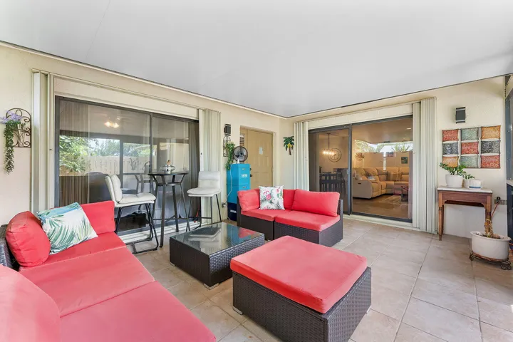 Spacious open-concept living area featuring coral upholstered seating with dark woven accent tables, tile flooring throughout, and expansive sliding glass doors opening to a covered patio